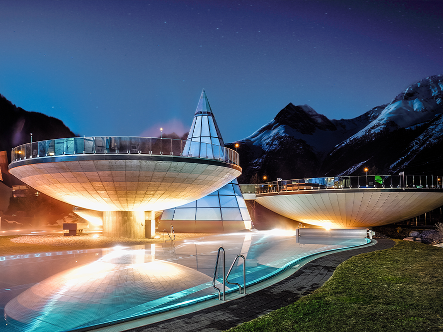 Am Fuße hoher Berge gelegen, erstrahlt der moderne Outdoor-Spa-Komplex des Thermenhotels mit seinen beleuchteten futuristischen Strukturen unter dem Nachthimmel und bietet einen ruhigen Rückzugsort für Ruhesuchende.