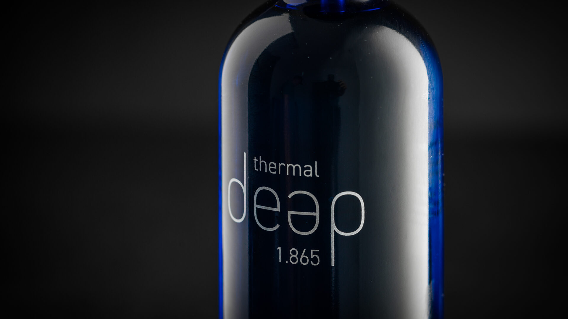Eine Nahaufnahme einer dunkelblauen Flasche mit der eleganten Aufschrift „Thermal Deep 1.865“ hebt sich vom tiefschwarzen Hintergrund ab.