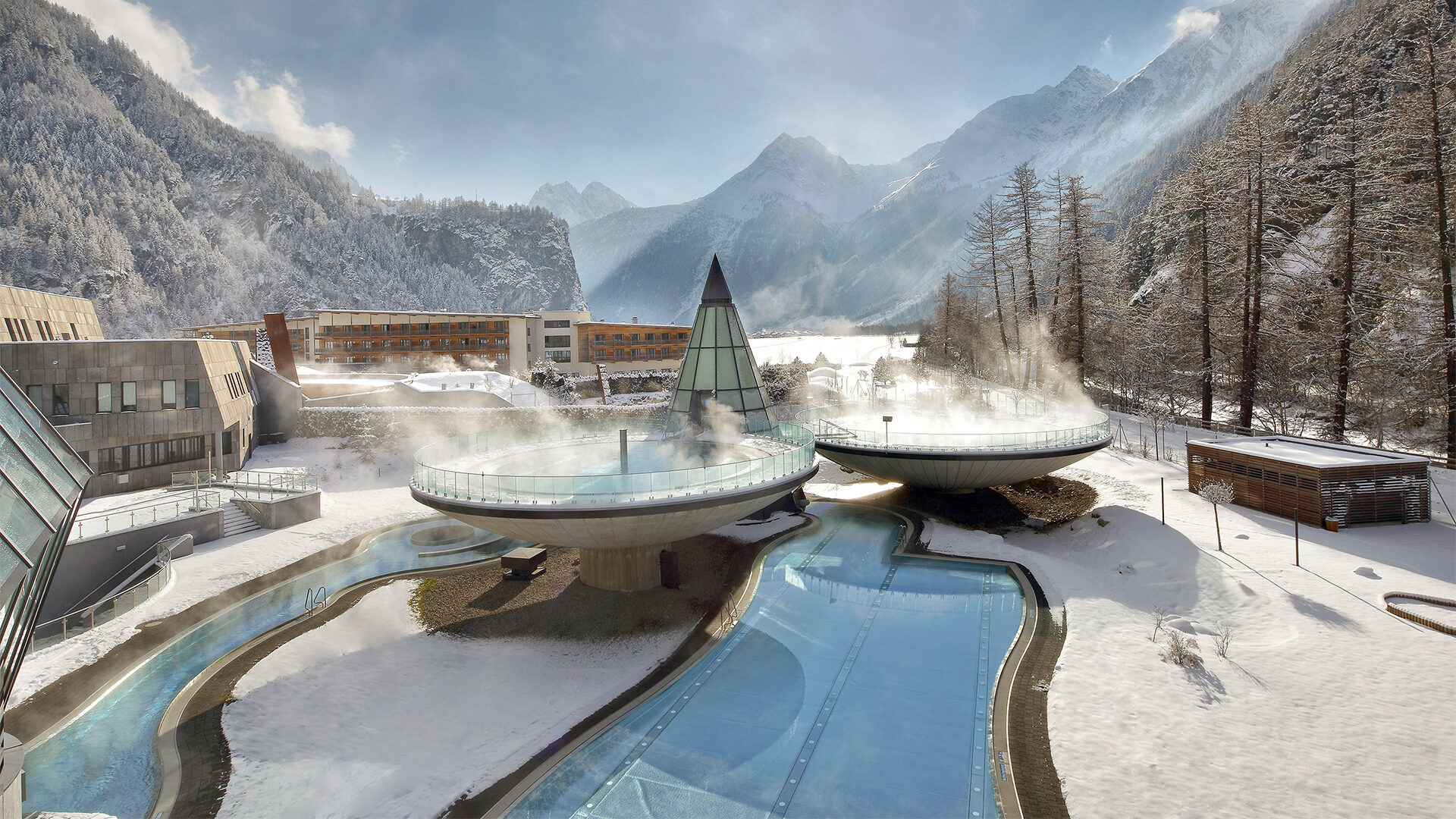 Snow-covered landscape with a modern spa resort, perfect for a winter holiday. Two steam-emitting circular structures are elevated above pools, surrounded by snowy trees and mountains under a blue sky.