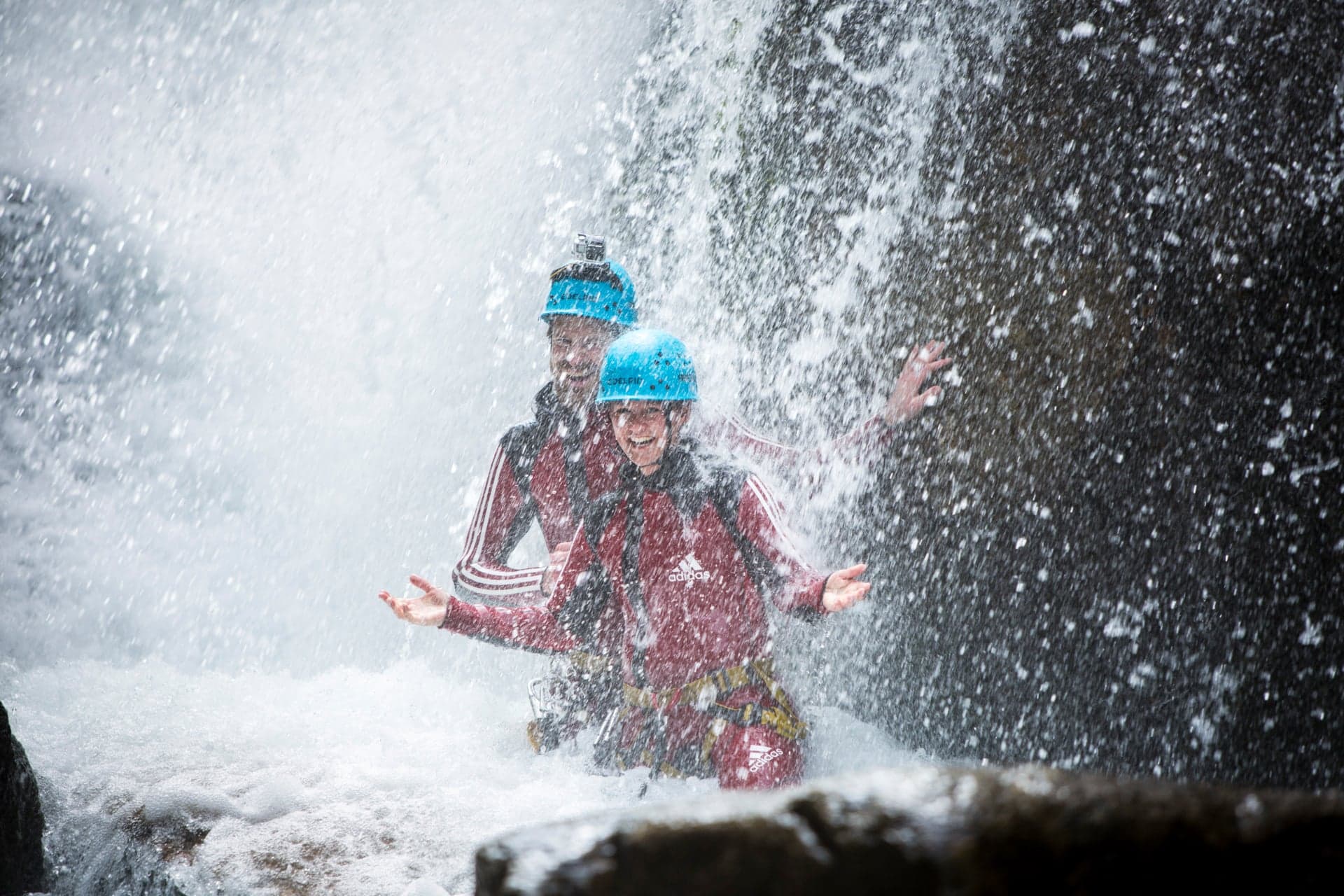 Zwei Menschen mit Helmen und roten Neoprenanzügen stehen lächelnd unter einem Wasserfall, während das Wasser um sie herum plätschert, und genießen eine abenteuerliche Variante ihres Thermenurlaubs.