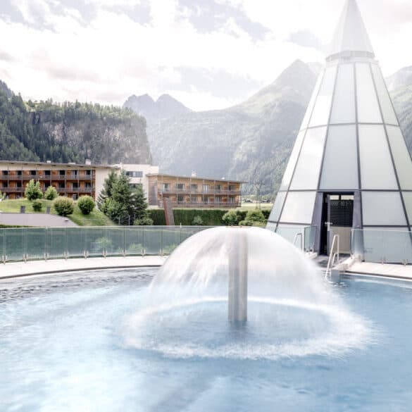 Фонтан на открытом воздухе в круглом бассейне с видом на горы. Рядом стоит современная стеклянная конструкция в форме конуса, на заднем плане — деревянное здание и зеленый ландшафт.