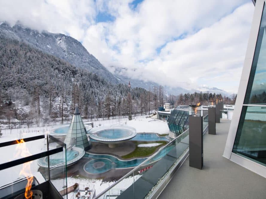 Snowy mountain landscape with modern glass buildings. Pool area outside with circular structures and trees in the background. Overcast sky above.