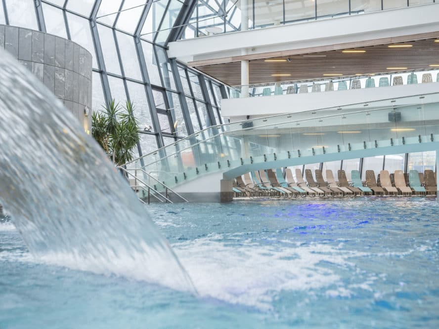 Indoor pool area with a waterfall feature, lounge chairs, and a glass ceiling. The space is airy and filled with natural light.