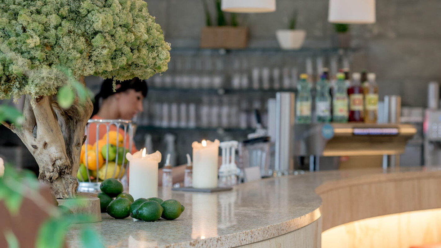 Le comptoir du café arbore une surface en marbre ornée de bougies gourmandes, d'une corbeille de fruits et d'une décoration en forme d'arbre. Des étagères garnies de bouteilles contribuent à l'ambiance, tandis qu'une silhouette floue en arrière-plan évoque l'atmosphère thermale.
