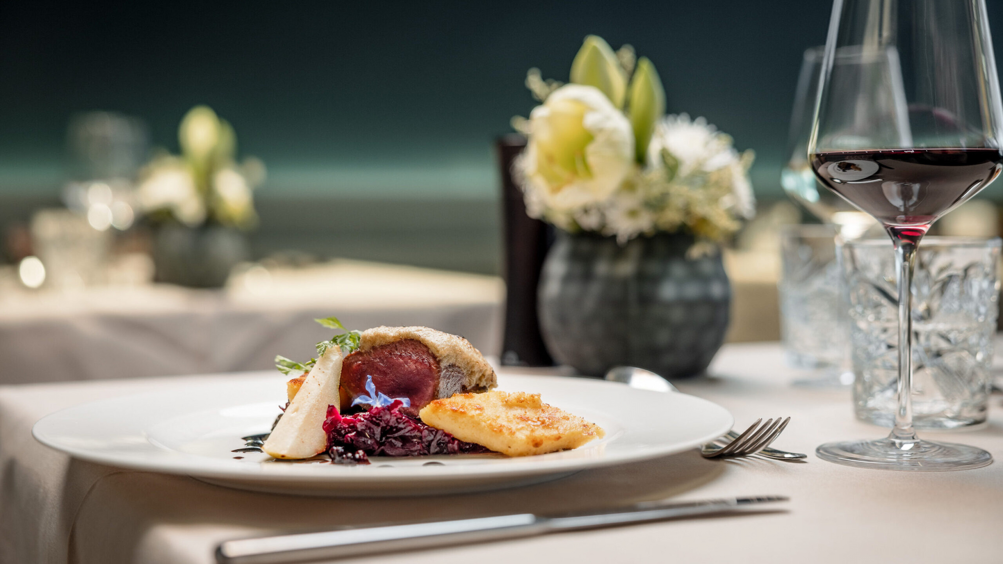 Un elegante ambiente da ristorante presenta un piatto di carne, cavolo rosso e patate elegantemente disposti su un piatto bianco. Un bicchiere di ottimo vino rosso e un centrotavola floreale aggiungono fascino allo sfondo, esaltando l'atmosfera sofisticata.