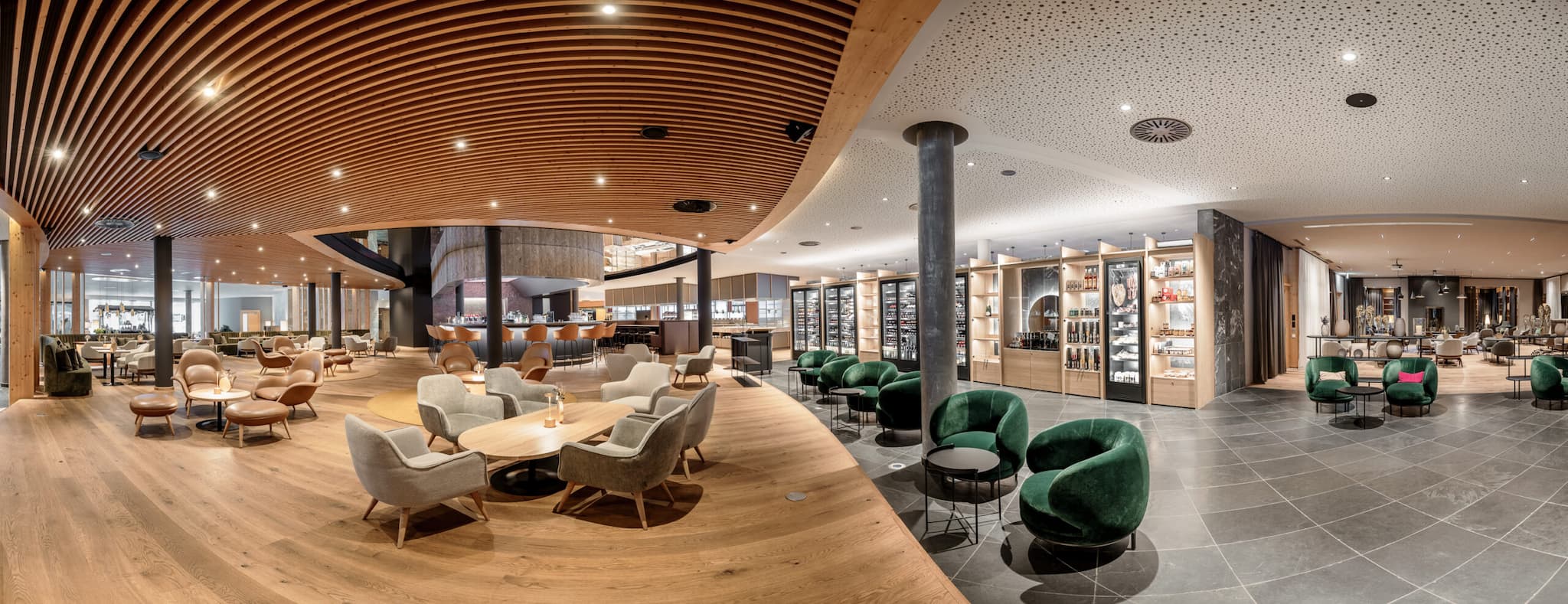 Geräumige, moderne Lounge mit Holzboden, verschiedenen Stühlen und Tischen. Regale präsentieren stolz verschiedene Gegenstände, die an einen perfekten Herbsturlaub erinnern. Die Decke ist mit Holzlamellen und Strahlern geschmückt und schafft eine einladende Atmosphäre zum Entspannen.