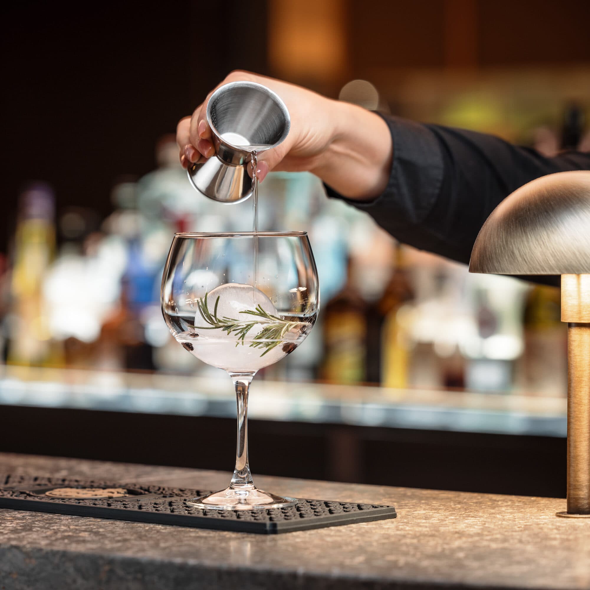 Al bancone di un bar affollato, che ricorda i ristoranti di lusso, una mano versa abilmente il liquido da un misurino in un grande bicchiere, abbracciando una sfera di ghiaccio e un rametto.