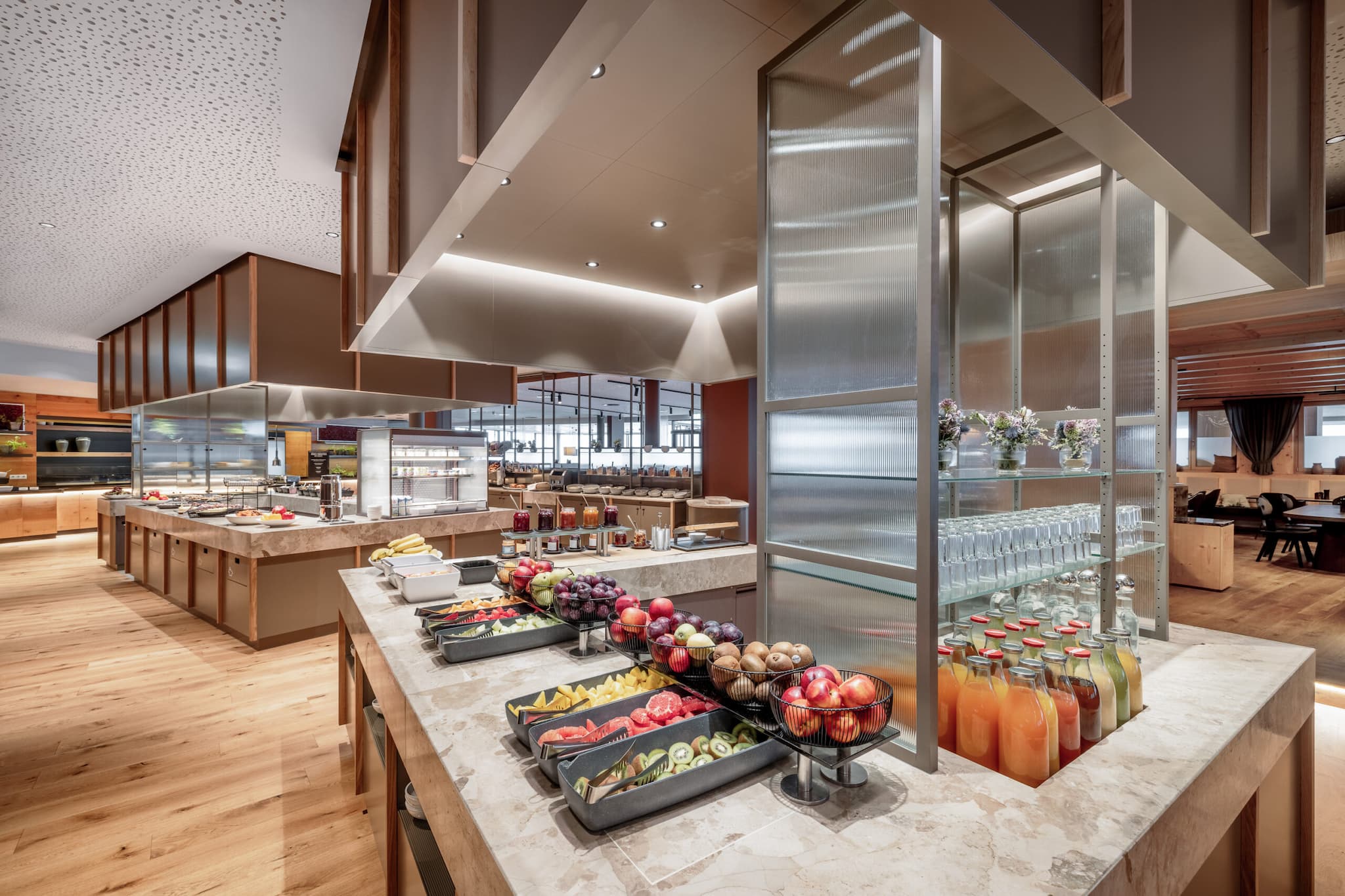 Genießen Sie ein herrliches kulinarisches Erlebnis mit einem Buffet mit verschiedenen Früchten, Gemüsesorten und frischen Säften, elegant angerichtet auf einer Marmortheke in einem modernen Ambiente mit Holzböden und stimmungsvoller Deckenbeleuchtung.