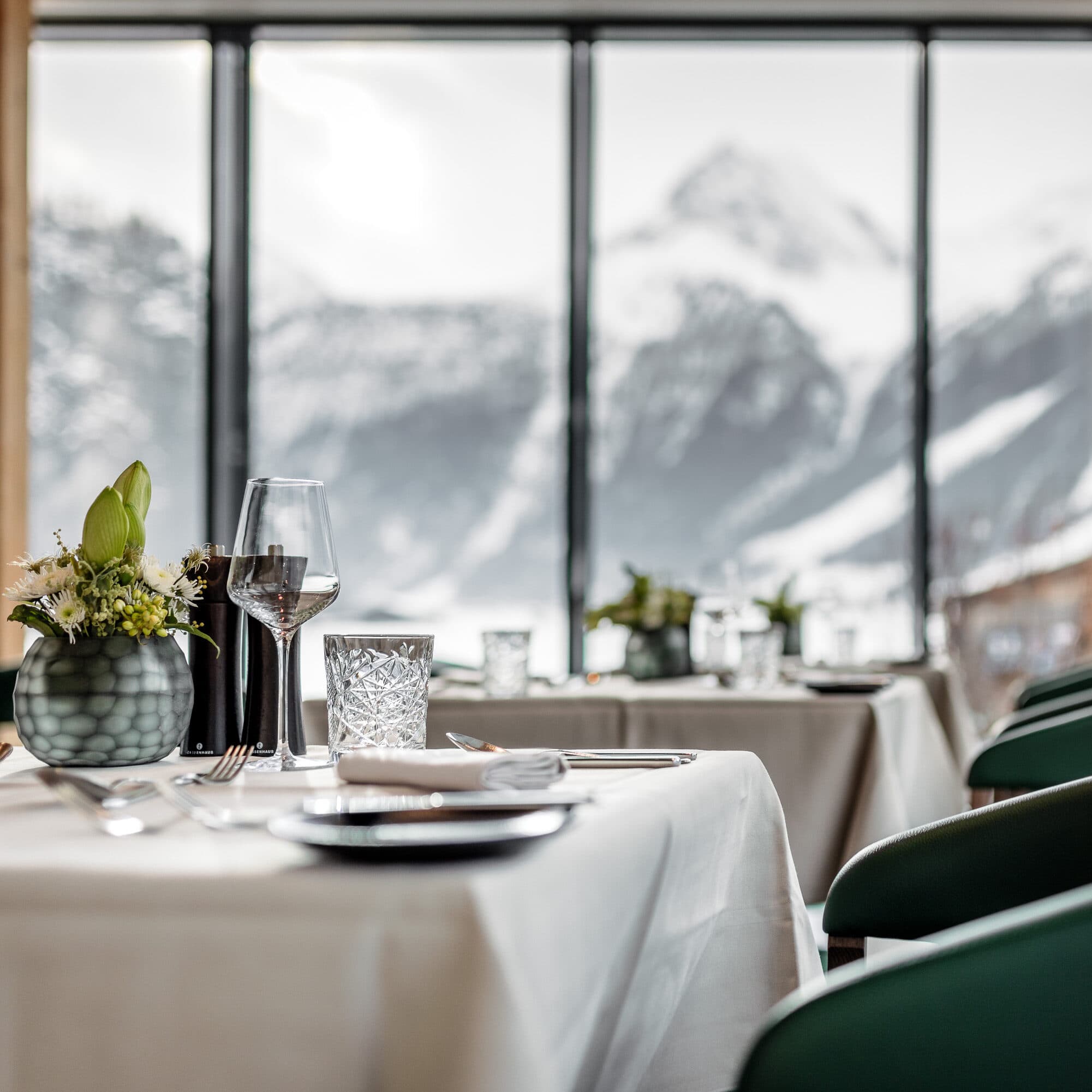 Elegante tavolo da pranzo apparecchiato con raffinati bicchieri e fiori freschi in un ristorante che offre viste mozzafiato sulle montagne innevate attraverso ampie finestre.