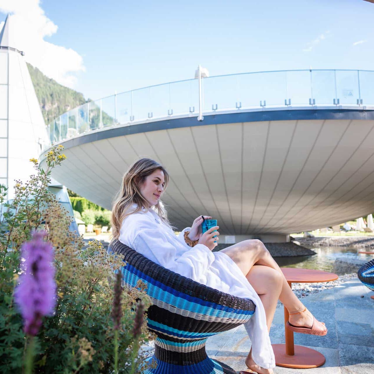 Une personne vêtue d'une robe blanche est assise sur une chaise tressée à l'extérieur, un téléphone à la main. Par cette journée ensoleillée, elle se trouve devant une grande structure culinaire ronde, entourée de plantes et de fleurs qui rehaussent l'atmosphère sereine.