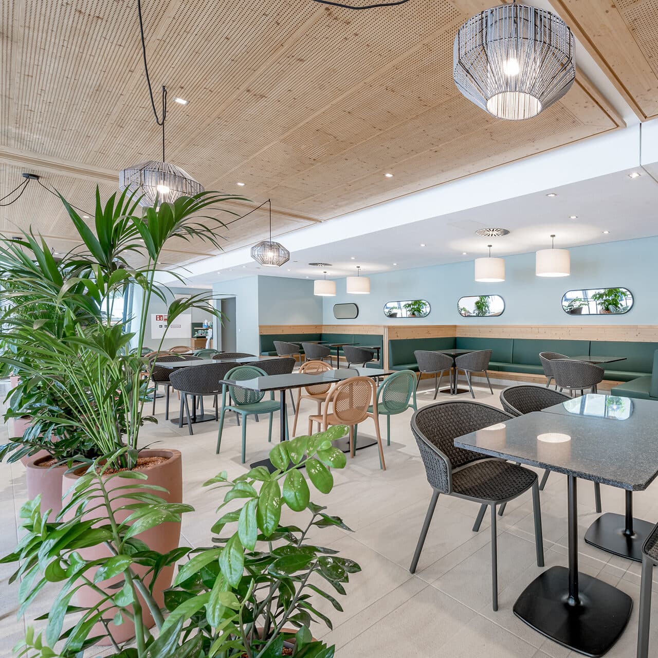 Intérieur de restaurant-café moderne avec plantes vertes, plafond en bois, suspensions rondes et chaises vertes et grises disposées autour des tables. De grandes fenêtres et des miroirs apportent lumière naturelle et reflets chaleureux.