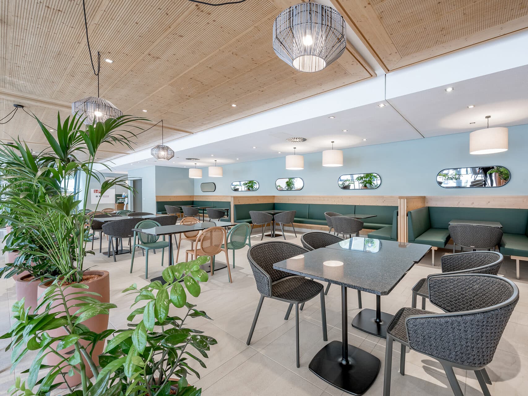 Intérieur de café moderne avec plafond en bois, chaises en osier et plantes en pot. Idéal pour les conférences, l'espace présente des murs bleus ornés de miroirs elliptiques surplombant des banquettes vertes rembourrées. Des suspensions diffusent une lumière douce, créant une atmosphère chaleureuse et accueillante pour les réunions.