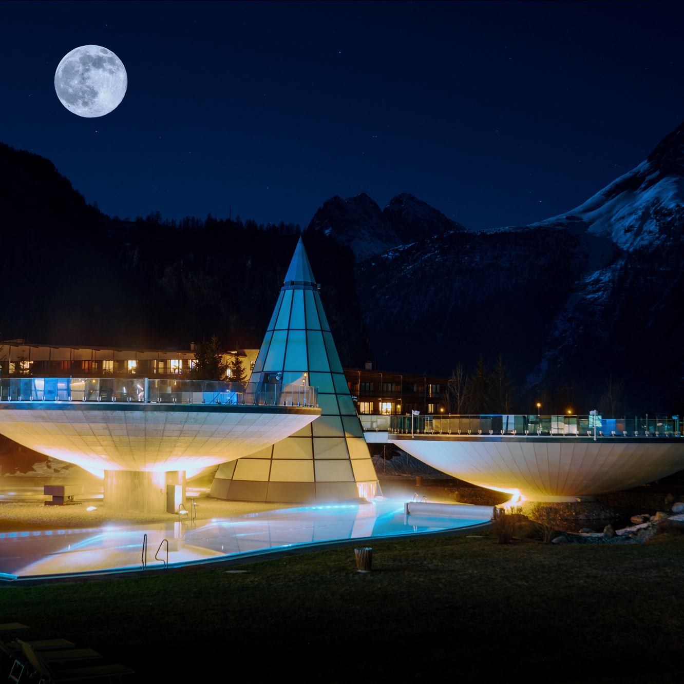 Una moderna piramide di vetro e due piscine rotonde illuminate si stagliano contro le montagne, sotto la luna piena di notte.