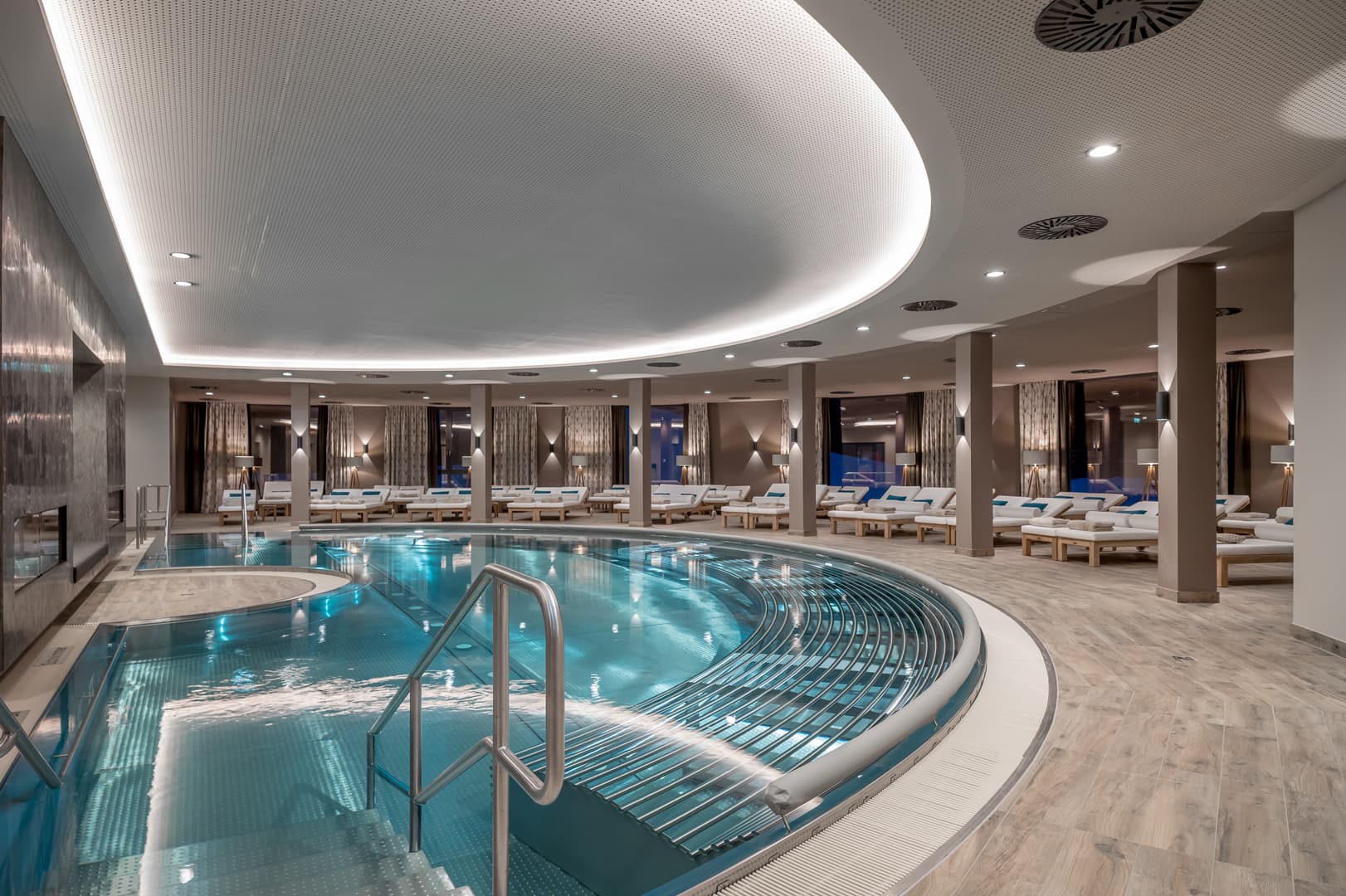 Entspannen Sie sich in unserem Indoor-Day-Spa mit modernem Pool, umgeben von Liegestühlen und geschwungenen Deckenleuchten. Das elegante Design schafft eine ruhige Atmosphäre, die perfekt zur Erholung einlädt.