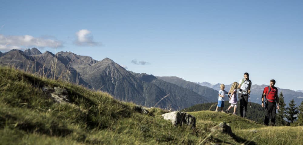 Eine vierköpfige Familie genießt ihren Herbsturlaub und wandert unter einem klaren, blauen Himmel auf einem grasbewachsenen Bergpfad mit malerischer Aussicht auf majestätische Gipfel im Hintergrund.