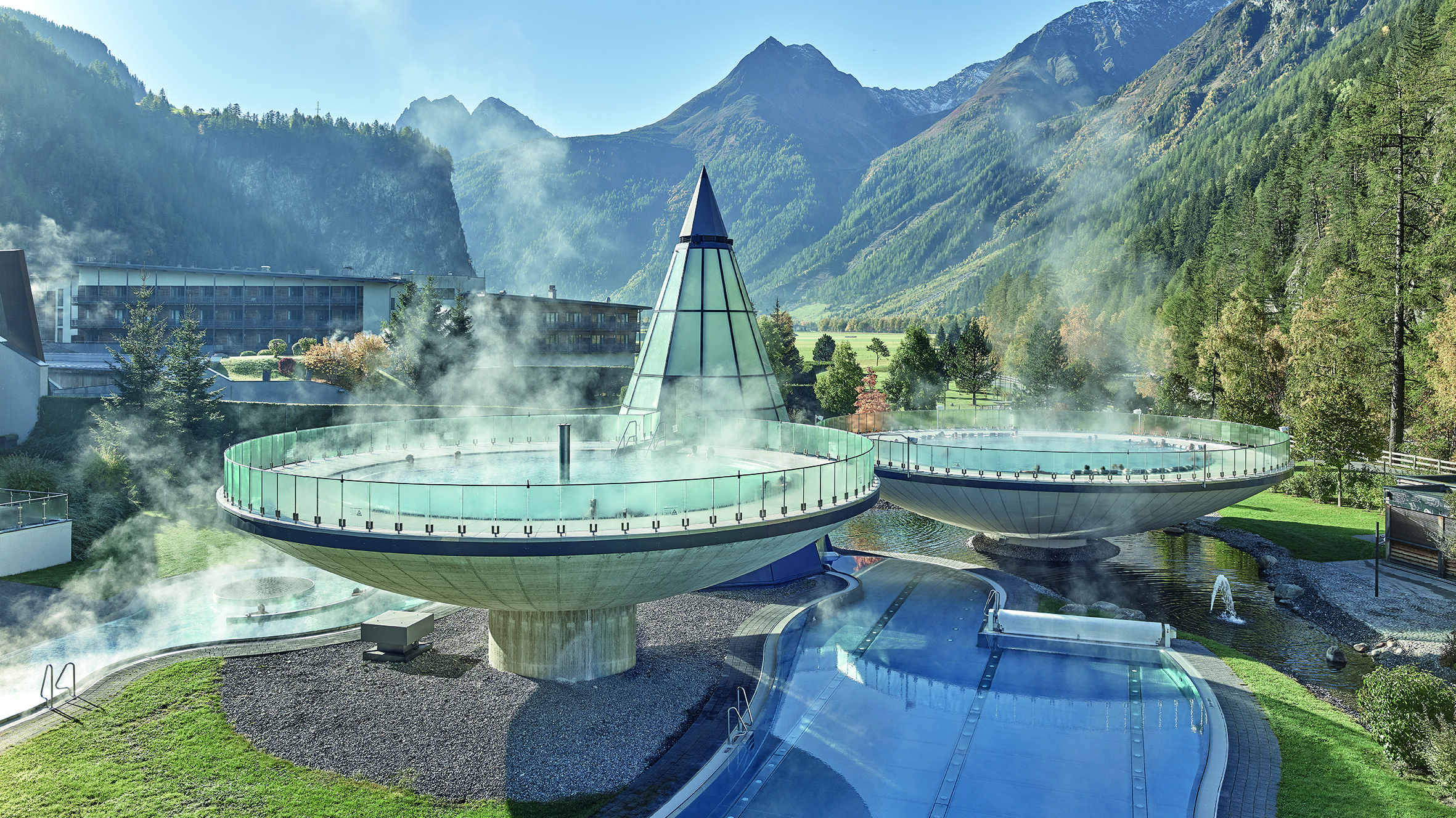 Due piscine termali circolari all'aperto emettono vapore accanto a una piramide di vetro, con uno sfondo di montagne e alberi: l'ambiente ideale per rilassanti vacanze autunnali.