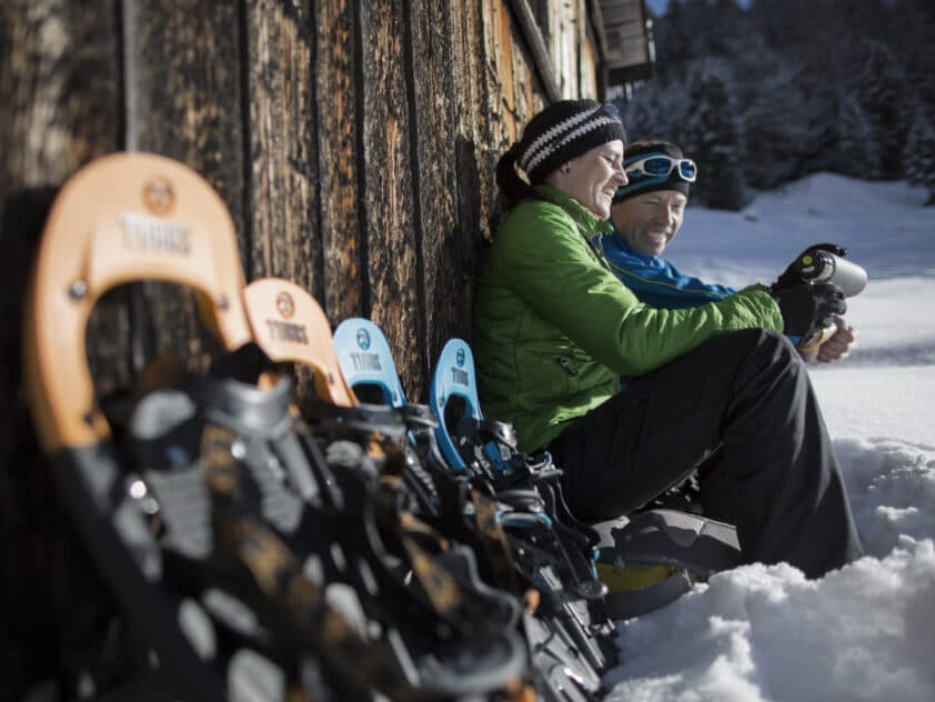 Zwei Personen in Winterkleidung sitzen während ihres Winterurlaubs lächelnd und mit Tassen in der Hand an einem Holzgebäude im Schnee, neben ihnen sind mehrere Paar Schneeschuhe aufgereiht.