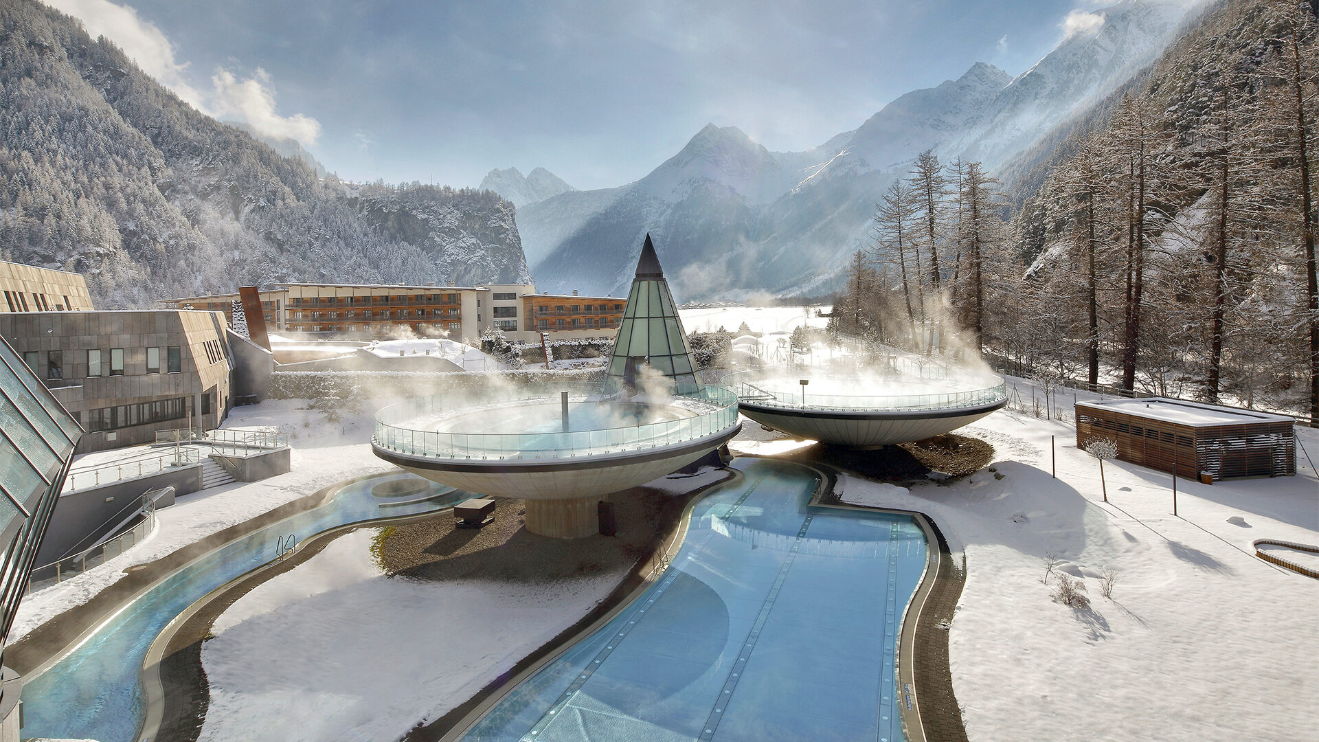 Il vapore si alza da grandi piscine circolari all'aperto in una località turistica di montagna innevata, circondata da edifici moderni e alberi innevati.