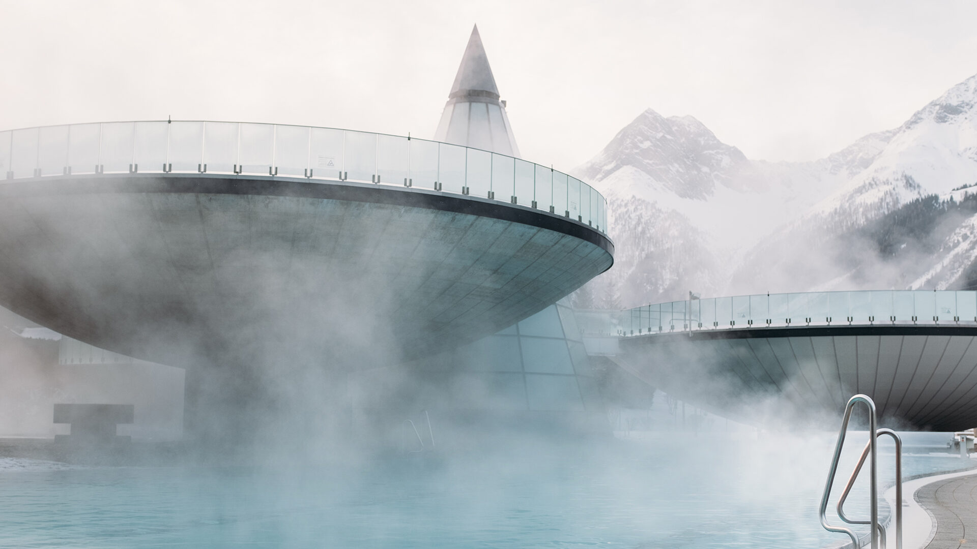 Futuristic circular building structures rise above a steamy outdoor pool with snowy mountains in the background.