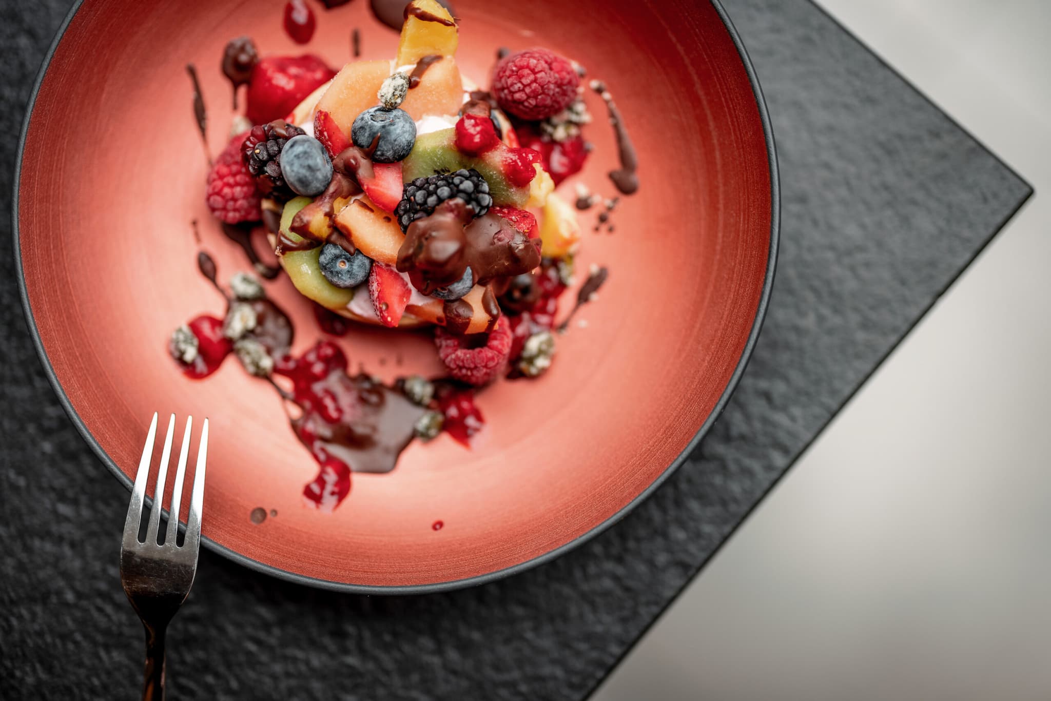 Auf einem roten Teller liegt ein farbenfrohes Kulinarik-Arrangement aus geschnittenem Obst, Beeren und Schokoladensoße; neben dem Teller liegt auf einer dunklen, strukturierten Oberfläche eine Gabel.