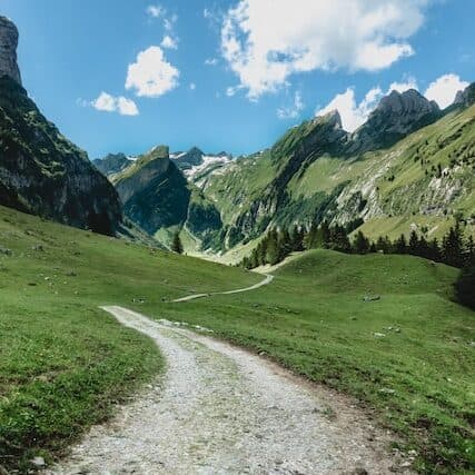 Un sentiero di ghiaia si snoda attraverso un prato verde circondato da montagne scoscese sotto un cielo parzialmente nuvoloso.