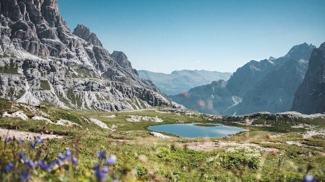 Un piccolo lago di montagna circondato da cime rocciose e prati erbosi sotto un cielo azzurro e terso, con fiori selvatici in primo piano.