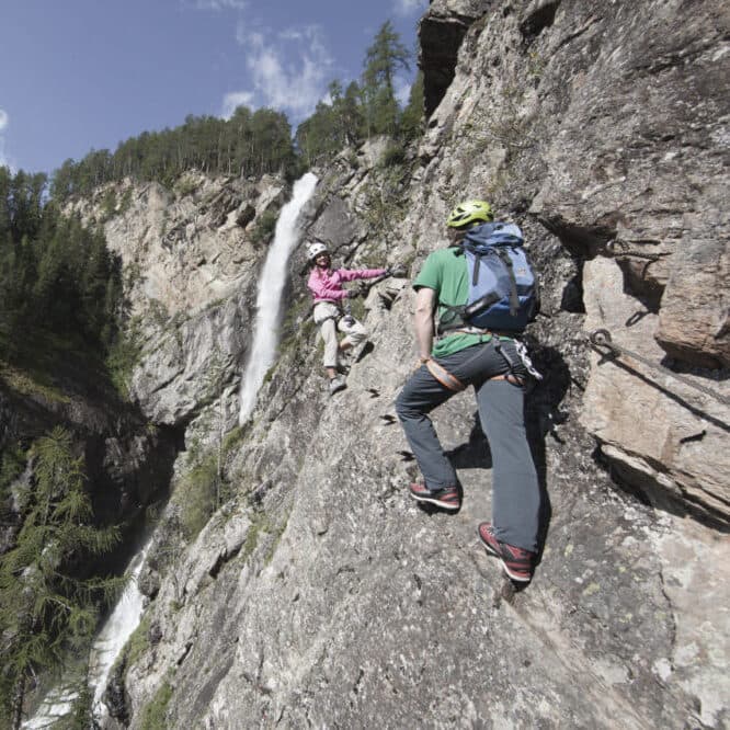 Due scalatori, muniti di caschi e imbracature, risalgono una ripida parete rocciosa accanto a una cascata, in una zona montuosa e boscosa.