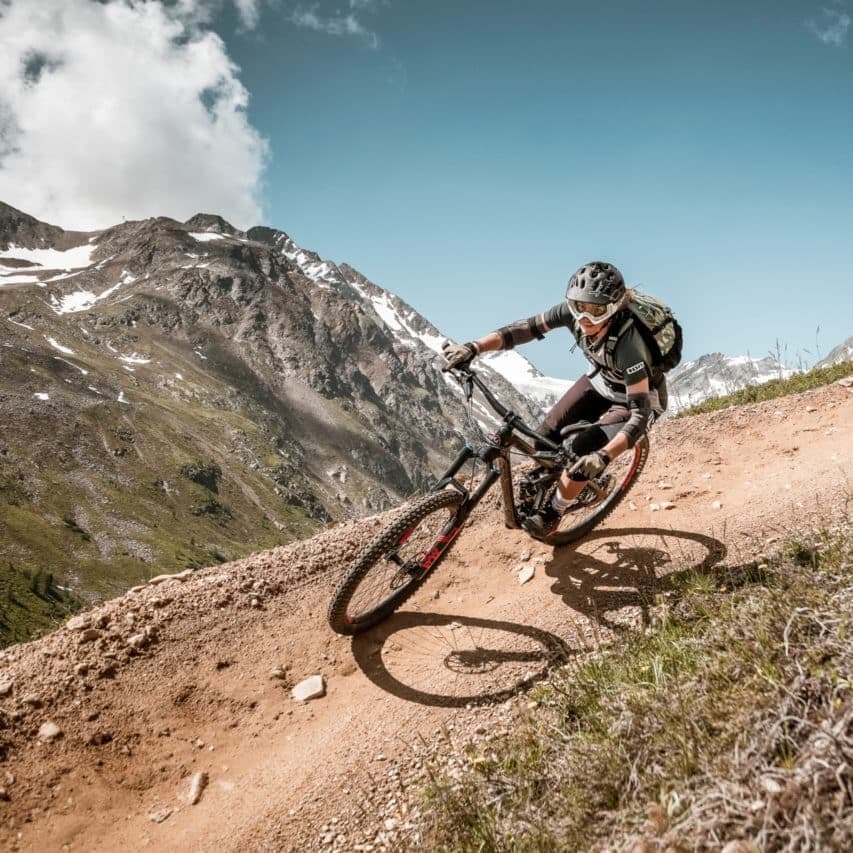 Un ciclista con casco e zaino percorre in mountain bike un sentiero sterrato con un terreno roccioso e montuoso sullo sfondo.