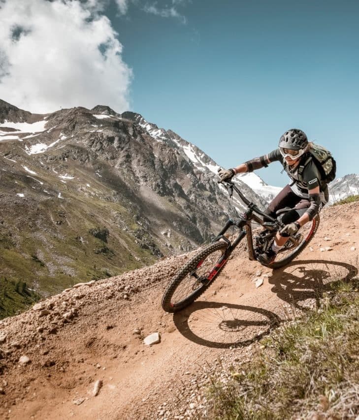 Una persona con casco e zaino percorre in mountain bike un sentiero sterrato, con montagne rocciose e un cielo azzurro sullo sfondo.
