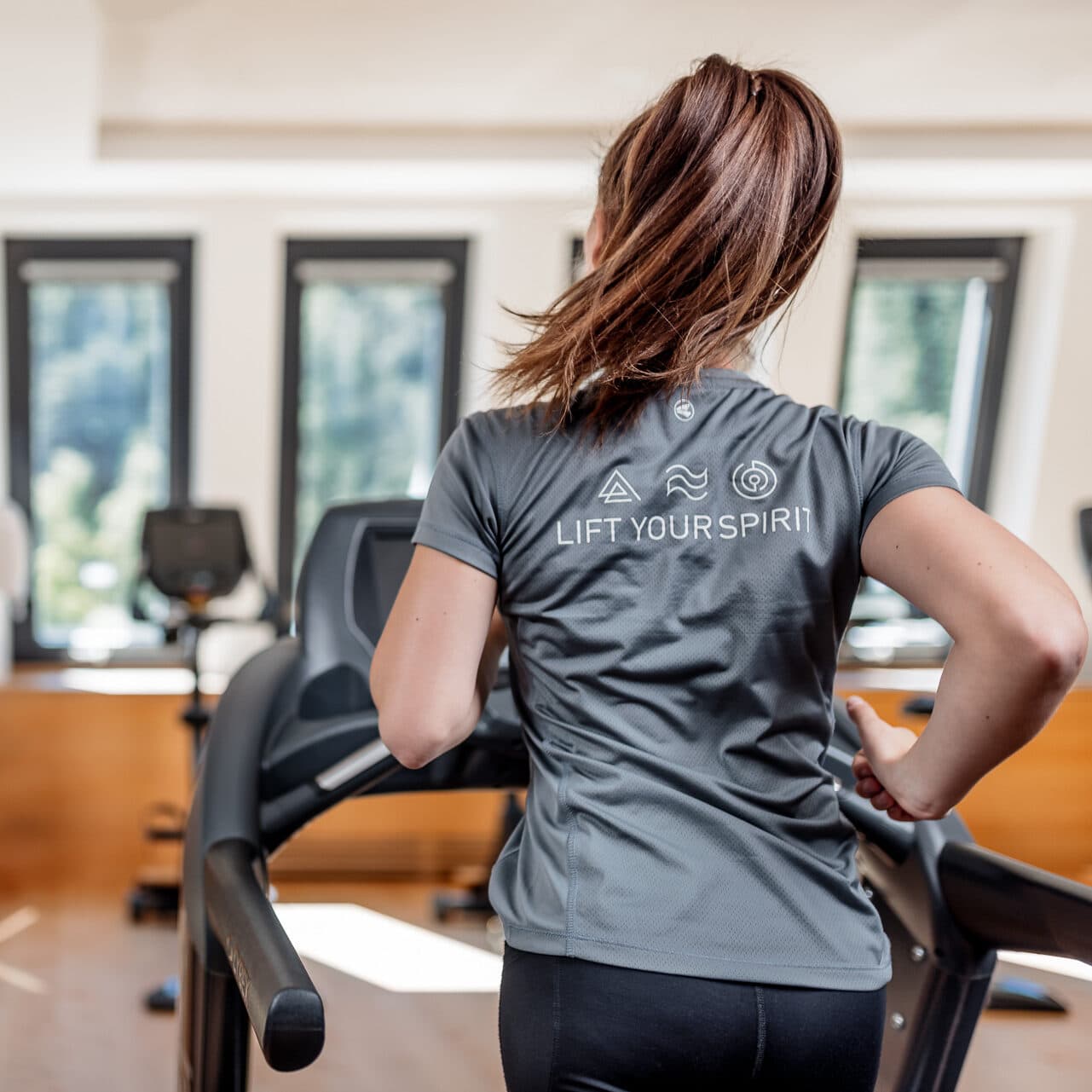 Eine Frau mit braunen Haaren läuft auf einem Laufband in einem hellen Fitnessstudio. Sie trägt ein graues Hemd mit der Aufschrift LIFT YOUR SPIRIT auf dem Rücken.