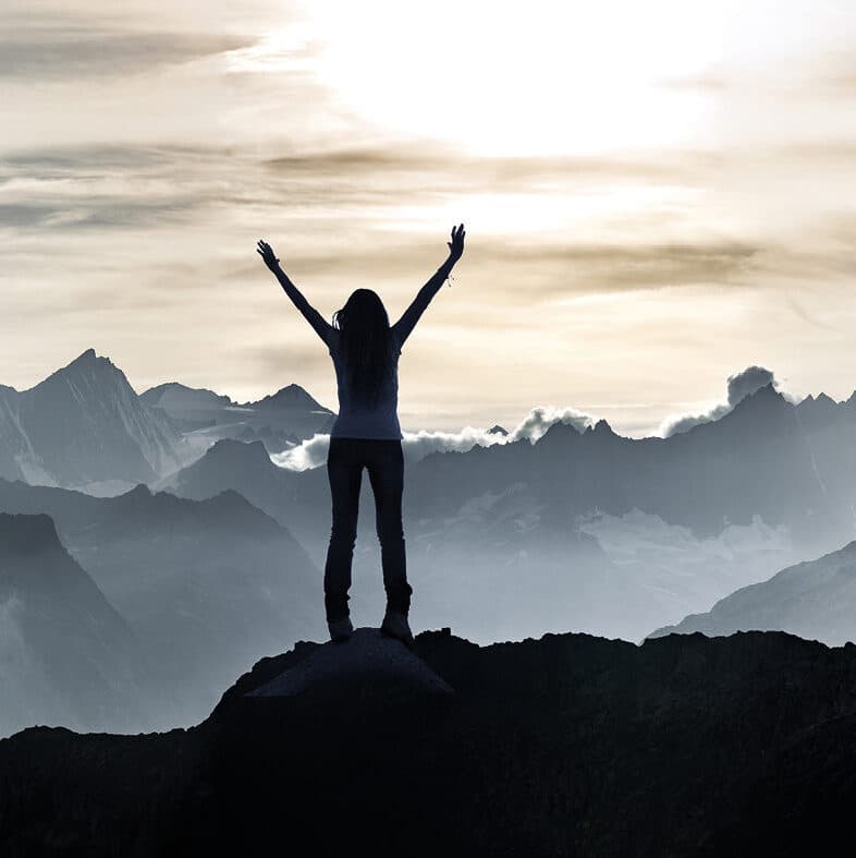 Eine Person steht mit erhobenen Armen auf einem Berggipfel und blickt bei Sonnenaufgang oder Sonnenuntergang unter einem bewölkten Himmel auf eine Gebirgskette.