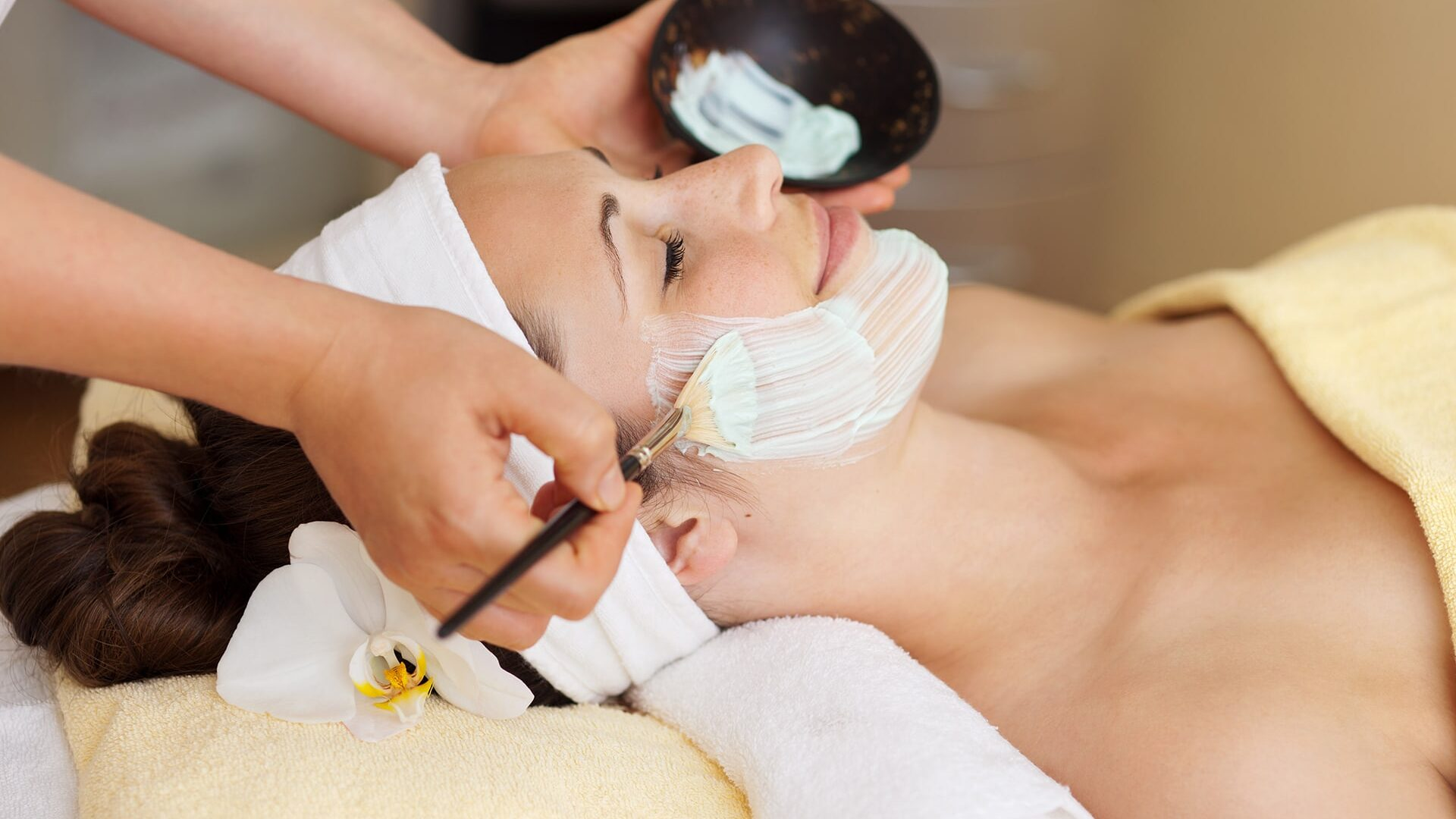 Eine Frau mit einem Stirnband und einer Blume im Haar erhält eine Gesichtsmaskenbehandlung mit einer Bürste, während sie auf einer Wellnessliege liegt.