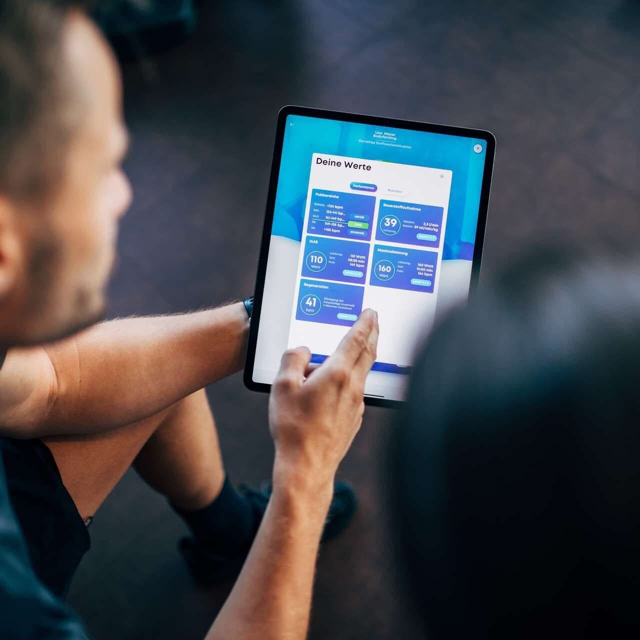 Eine Person hält ein Tablet, auf dem ein Gesundheits- oder Fitness-Dashboard in deutscher Sprache angezeigt wird, während eine andere Person in der Nähe sitzt.