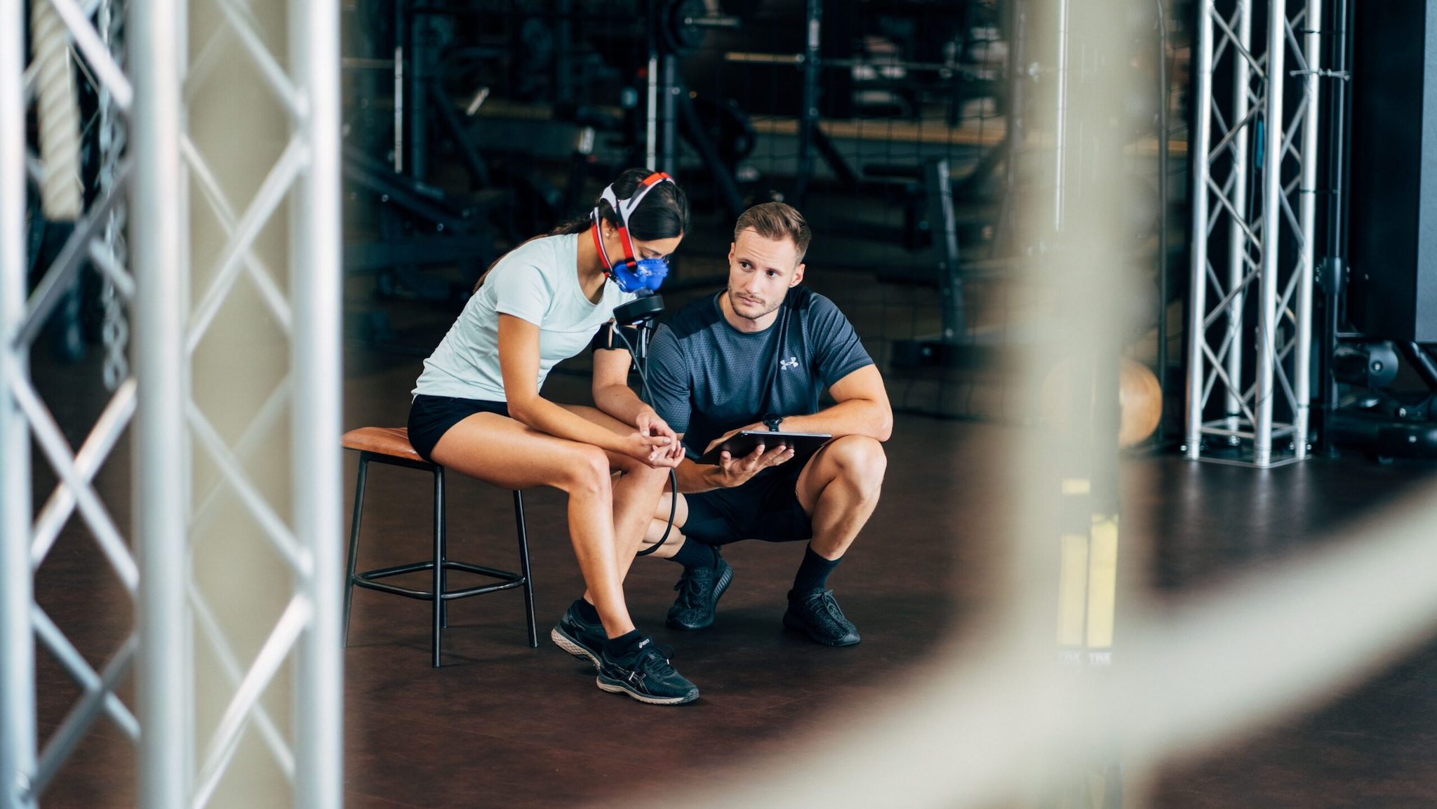 Eine Frau mit Sportkleidung und Atemmaske sitzt auf einer Bank, während ein Mann in Sportkleidung neben ihr kniet. Beide schauen in einem Fitnessstudio auf ein Tablet.