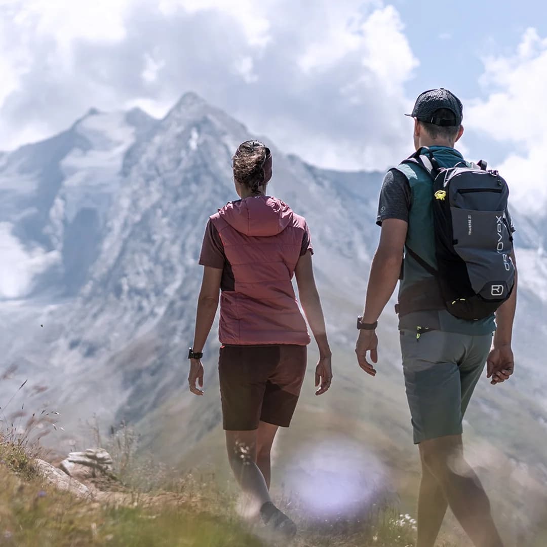 Zwei Personen in Outdoor-Kleidung und mit Rucksäcken wandern auf einem grasbewachsenen Weg in Richtung eines schneebedeckten Berges unter einem teilweise bewölkten Himmel.