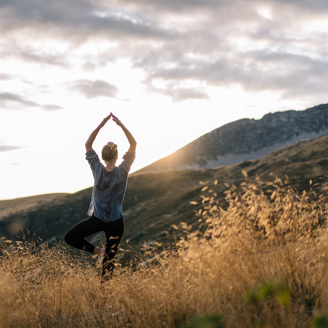 Eine Person steht in einer Yogastellung auf einem Bein in einem grasbewachsenen Feld mit Bergen und einer untergehenden Sonne im Hintergrund.