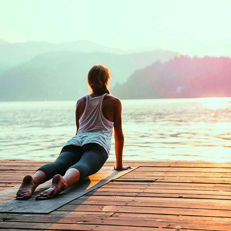 Person, die auf einer Matte auf einem hölzernen Steg an einem See mit Blick auf das Wasser bei Sonnenaufgang oder Sonnenuntergang Yoga übt, mit Bergen im Hintergrund.