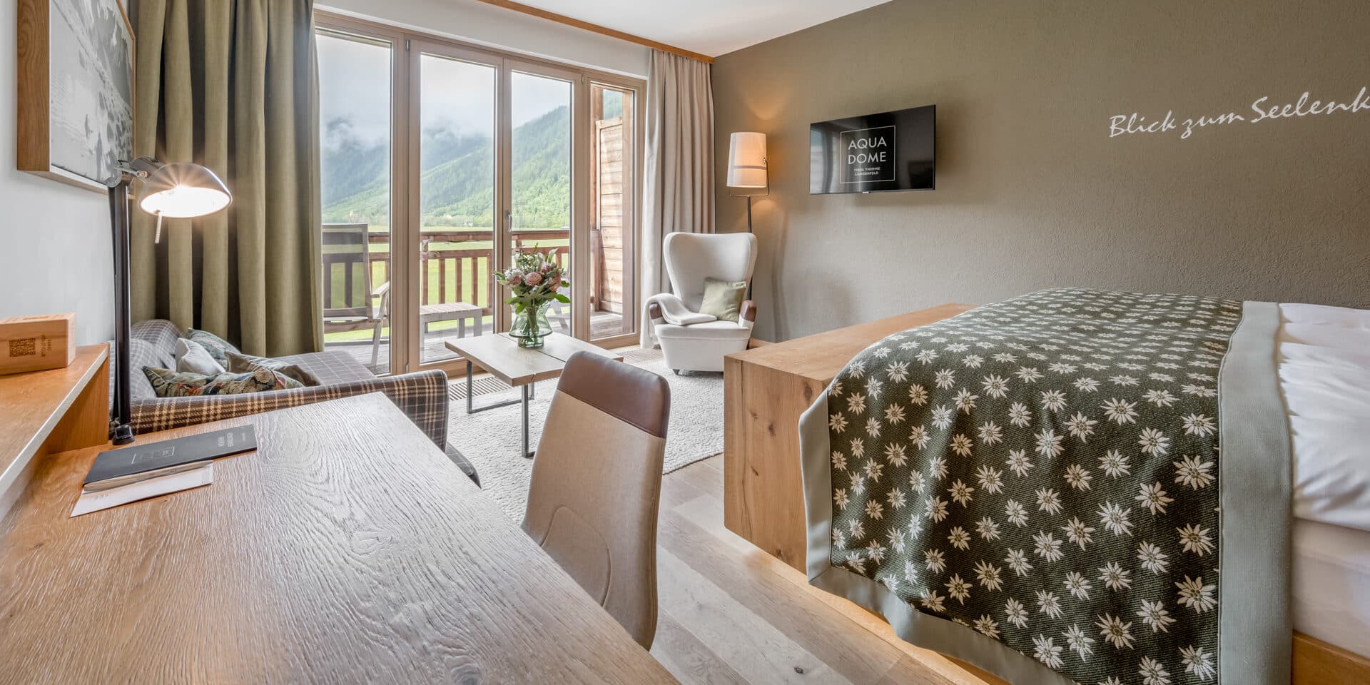 Modernes Hotelzimmer mit Bett, Schreibtisch, Sessel, Sofa, TV und Balkon mit Blick auf eine malerische Berglandschaft durch große Fenster.