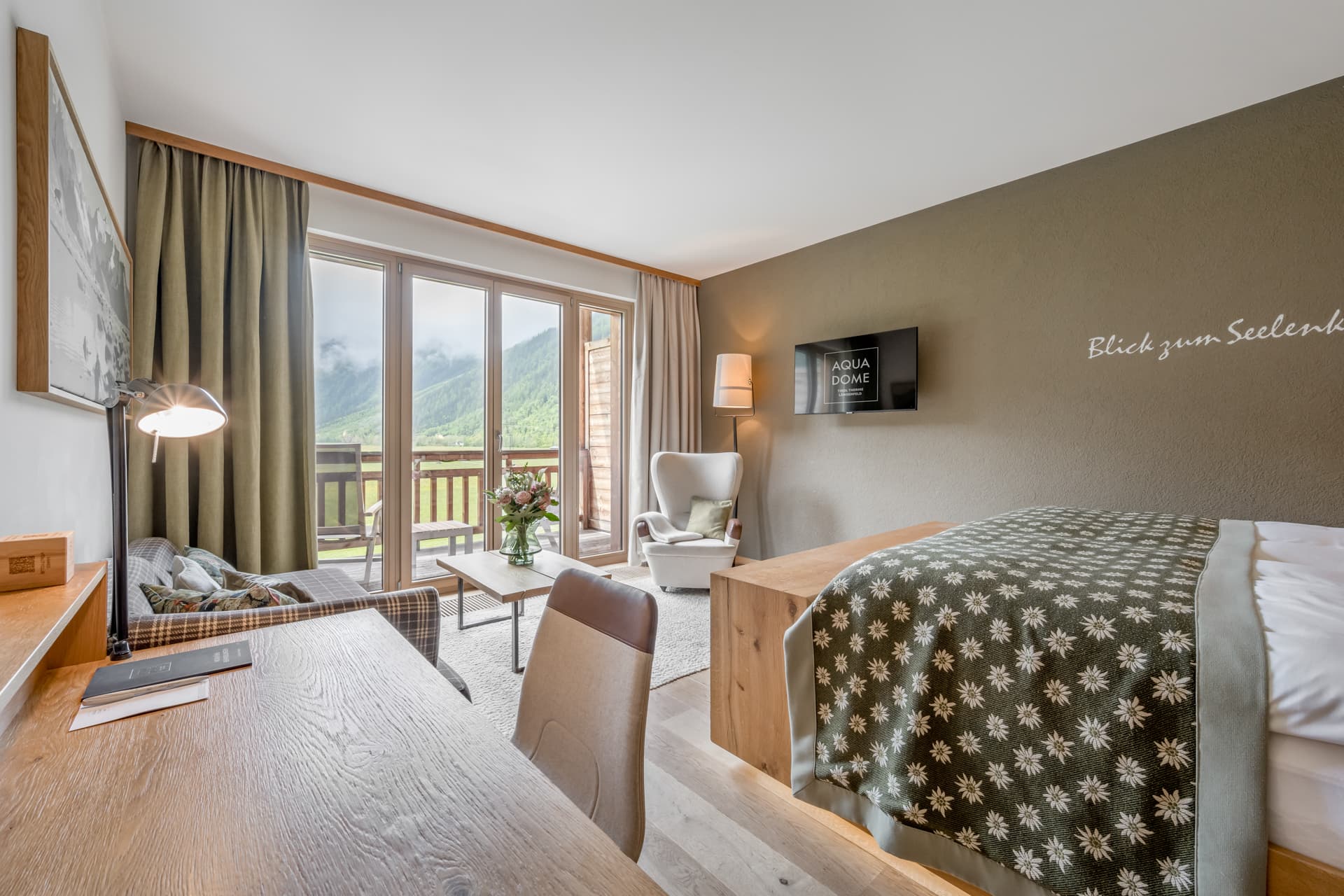 Современный гостиничный номер с двуспальной кроватью, письменным столом, креслом и диваном, с большими окнами и балконом с видом на горный пейзаж.