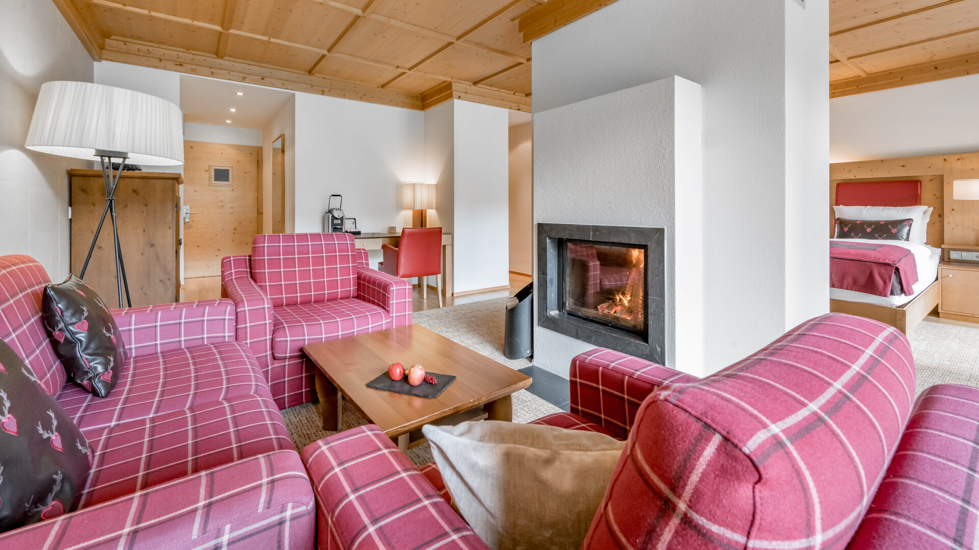 Une suite d'hôtel confortable avec des fauteuils à carreaux rouges, une table basse en bois, une cheminée et un lit visible à l'arrière-plan.