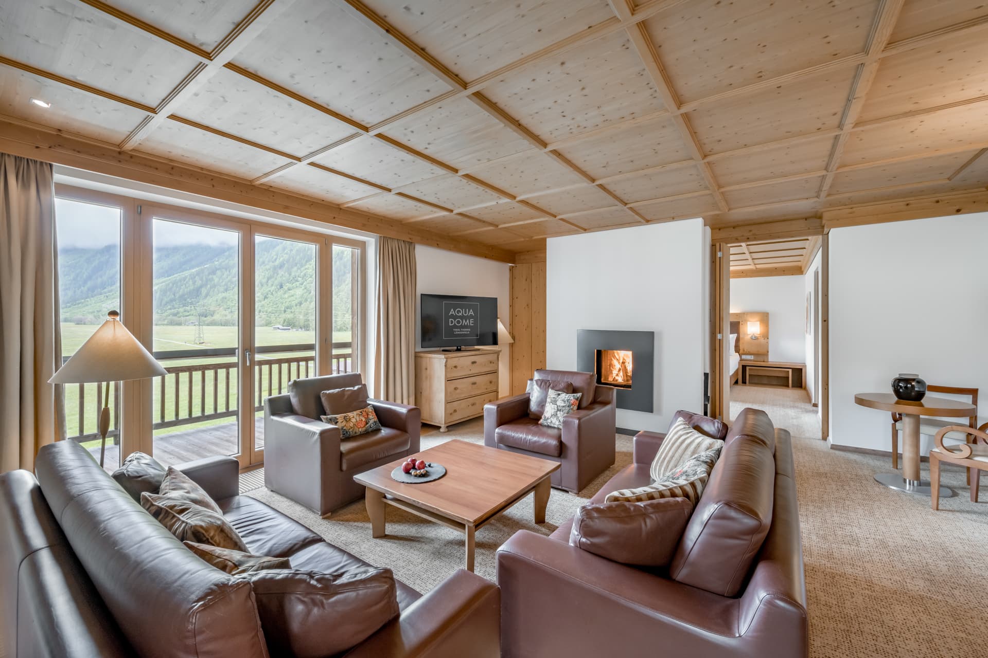 Просторная гостиная с кожаными диванами, деревянным столом, большими окнами с видом на горы, камином, телевизором и обеденной зоной в современном интерьере шале.