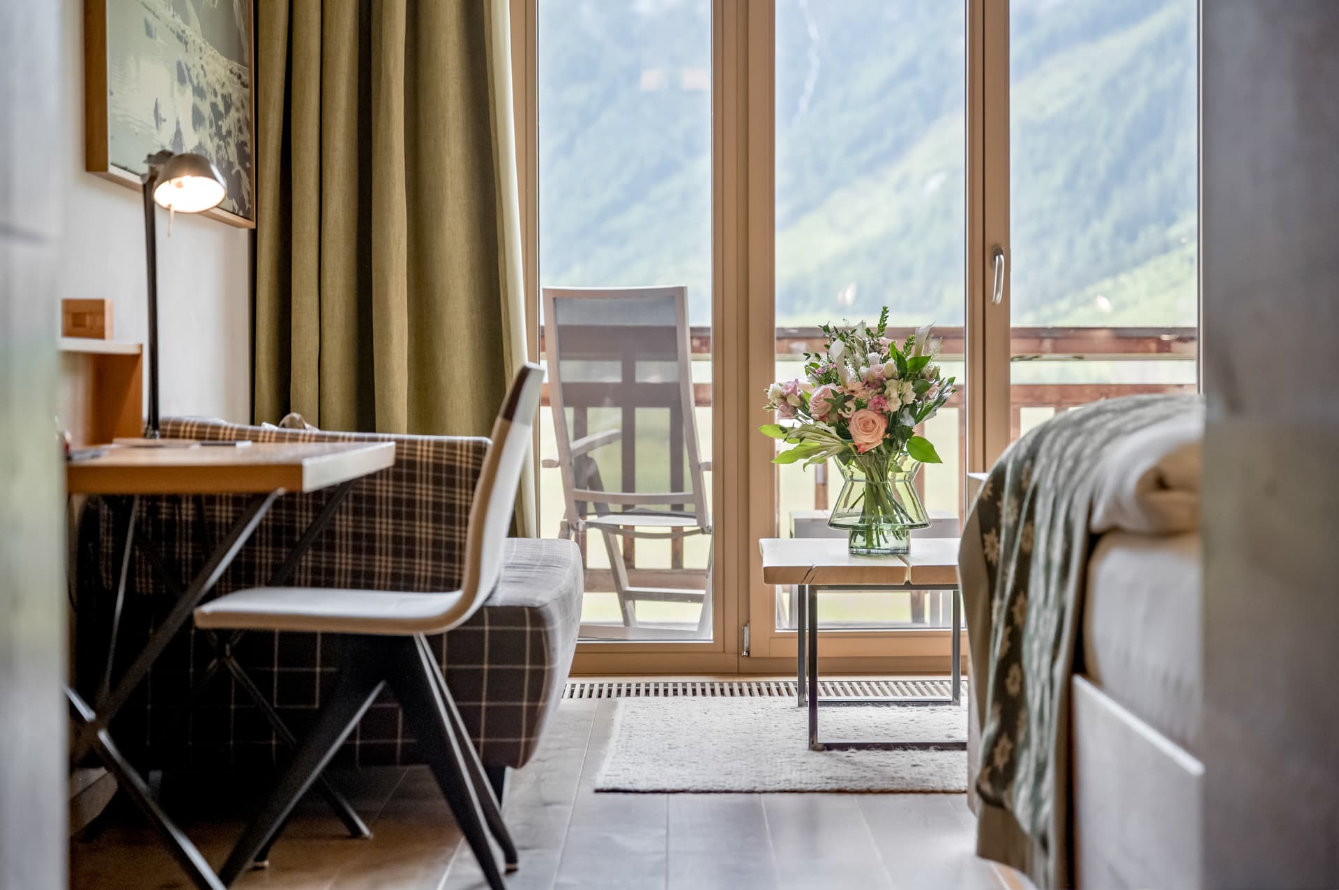 Гостиничный номер с письменным столом, стулом и кроватью, с вазой с цветами на столе перед стеклянными дверями, выходящими на балкон, за которым видны горы.