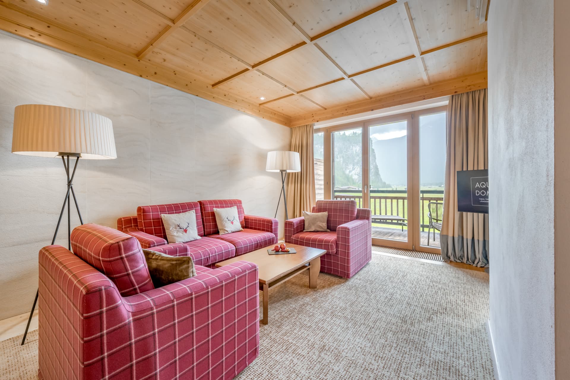 Гостиная с красными клетчатыми диванами, деревянным журнальным столиком, двумя торшерами, большими окнами с видом на улицу и бежевым ковровым покрытием.