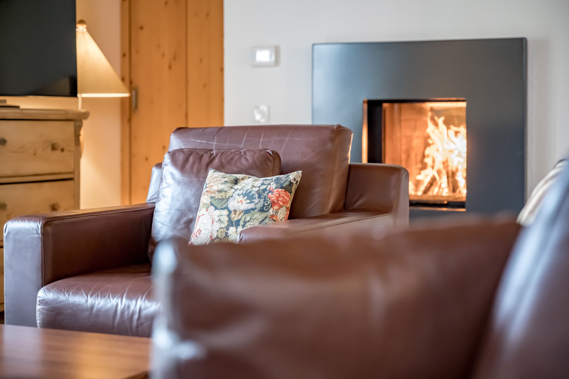 Коричневое кожаное кресло с цветочной подушкой стоит перед современным камином с зажженным огнем, рядом с деревянным шкафом и зажженной лампой.