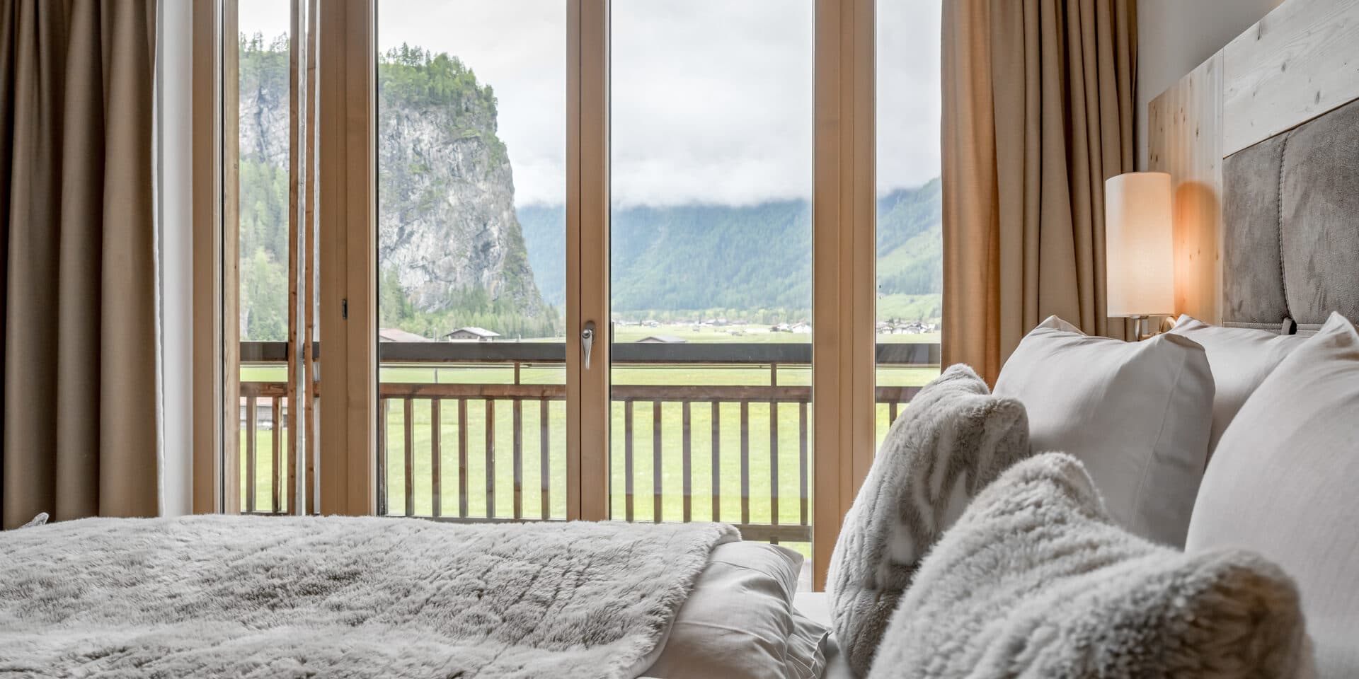 Ein gemütliches Schlafzimmer mit einem großen Bett und weichen Decken steht vor Glastüren, die auf einen Balkon mit Blick auf die Berge und das Tal führen.