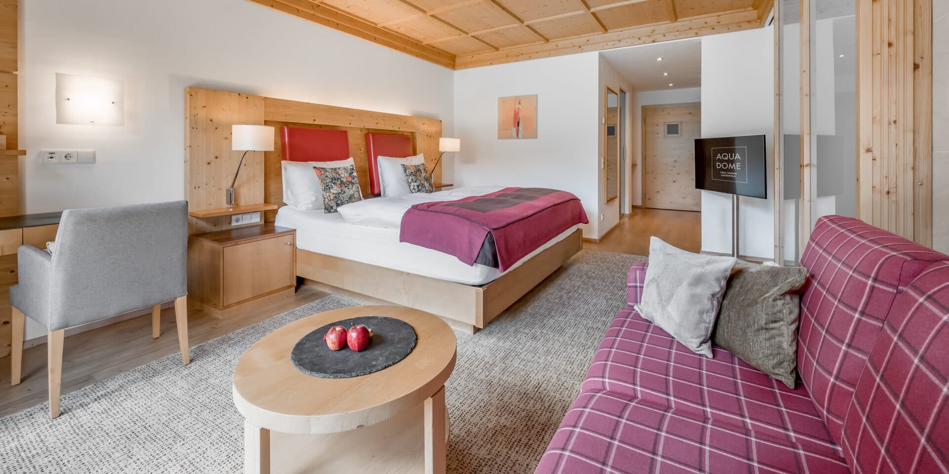 Modernes Hotelzimmer mit Doppelbett, Schreibtisch, Sofa, Flachbildfernseher, Holzakzenten und einem runden Couchtisch mit Äpfeln auf einer Schieferplatte.
