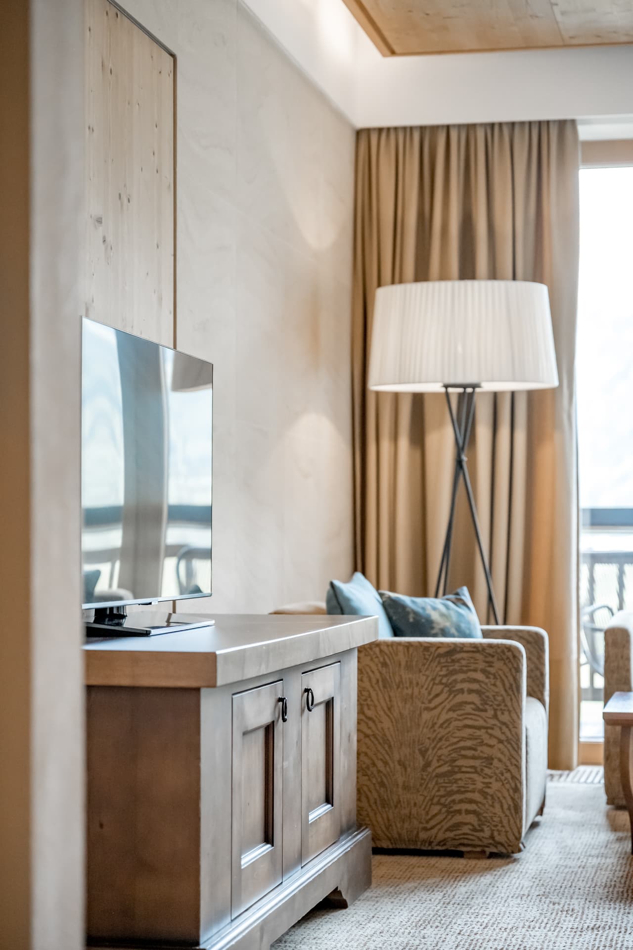 Современная гостиная с телевизором на деревянной тумбе, креслом с подушками, торшером и большими шторами, закрывающими окно.