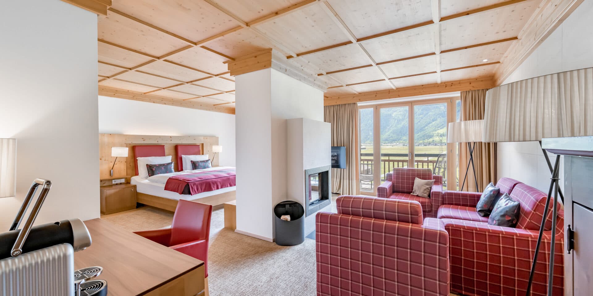Eine Hotelsuite mit einem Doppelbett, einem Schreibtisch und einer Sitzecke mit karierten Sesseln, die durch eine Teilwand abgetrennt sind, mit großen Fenstern und Blick auf die Berge.