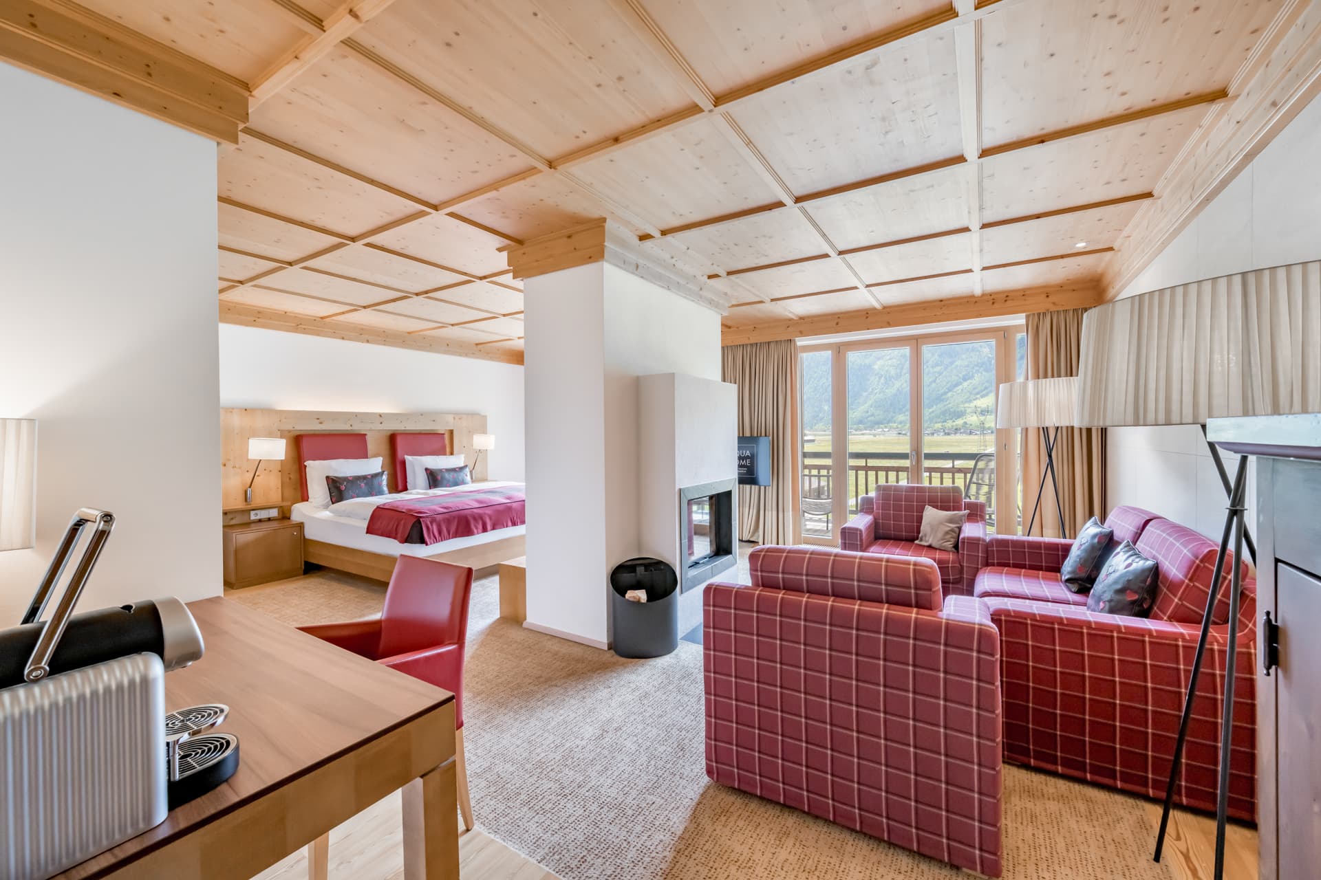 Современный гостиничный люкс с деревянным потолком, открытой планировкой, двуспальной кроватью, зоной отдыха с красными креслами и диваном, письменным столом и большими окнами с видом на горы.