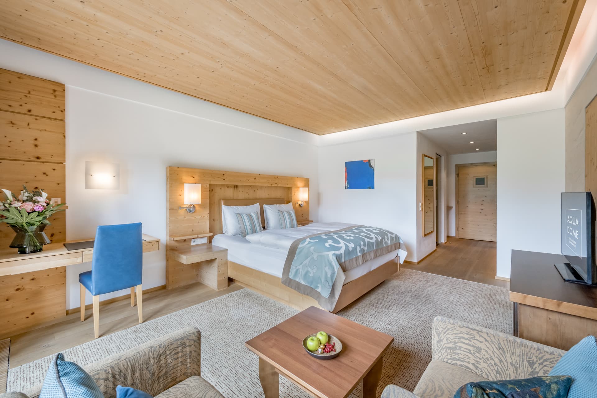 Современный гостиничный номер с деревянной мебелью, двуспальной кроватью, письменным столом со стулом, диваном, журнальным столиком с фруктами и телевизором. Номер хорошо освещен и оформлен в нейтральных и синих тонах.